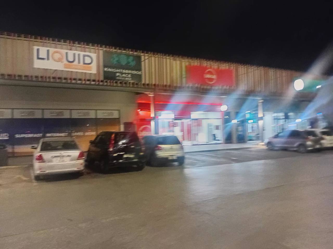 Illuminated retail and service storefronts with parked vehicles at Centro Mall in Kabulonga