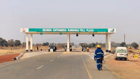 Reuben Chitandika Kamanga Toll Plaza on a Zambian highway, with a road worker in blue overalls walking past the toll booths and a minibus queued at the gate