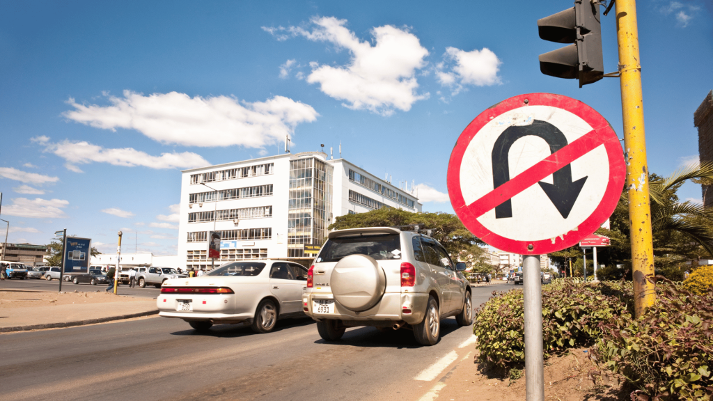 A No U-Turn traffic sign stands over a road in Zambia, with cars heading toward a commercial building under a blue sky