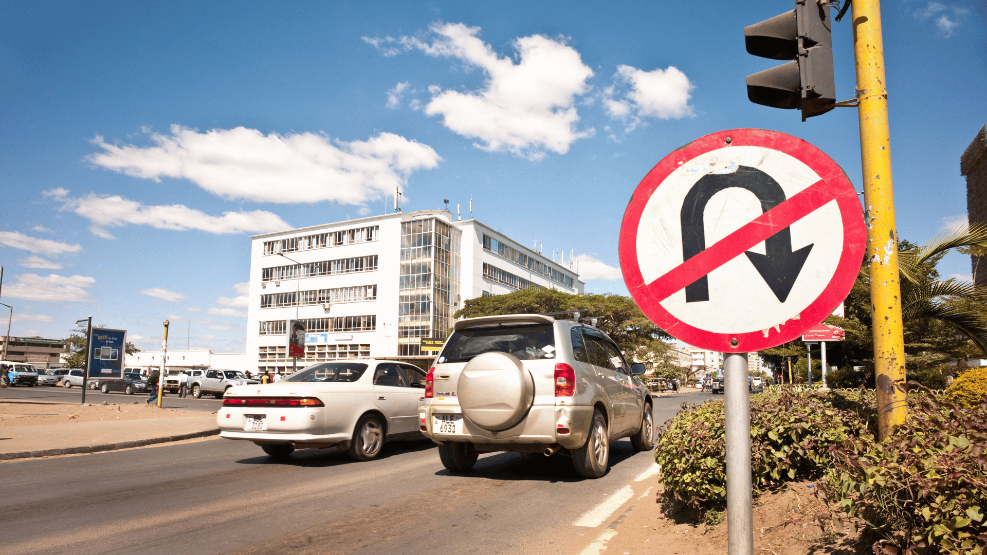 A No U-Turn traffic sign stands over a road in Zambia, with cars heading toward a commercial building under a blue sky