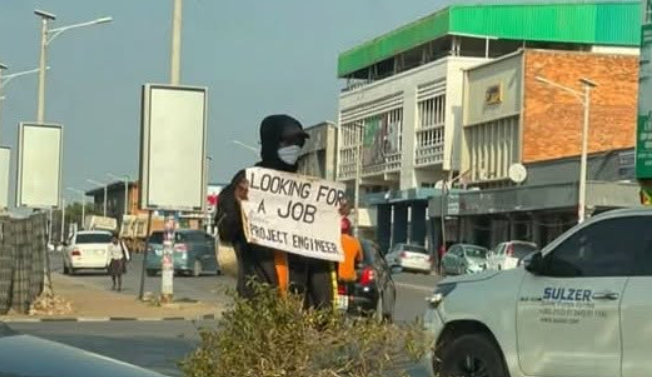 Job seeker in a dark hoodie and face mask holding a cardboard sign reading LOOKING FOR A JOB - PROJECT ENGINEER at an urban intersection.