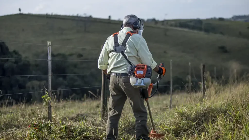 Farm worker wearing a harness, ear defenders, visor and gloves clears long grass along a wire fence line with a petrol brushcutter