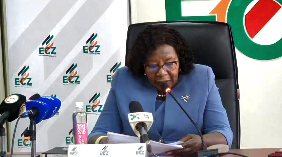 ECZ Chairperson Mwangala Zaloumis reads from papers at a media briefing with microphones and Electoral Commission of Zambia branding.