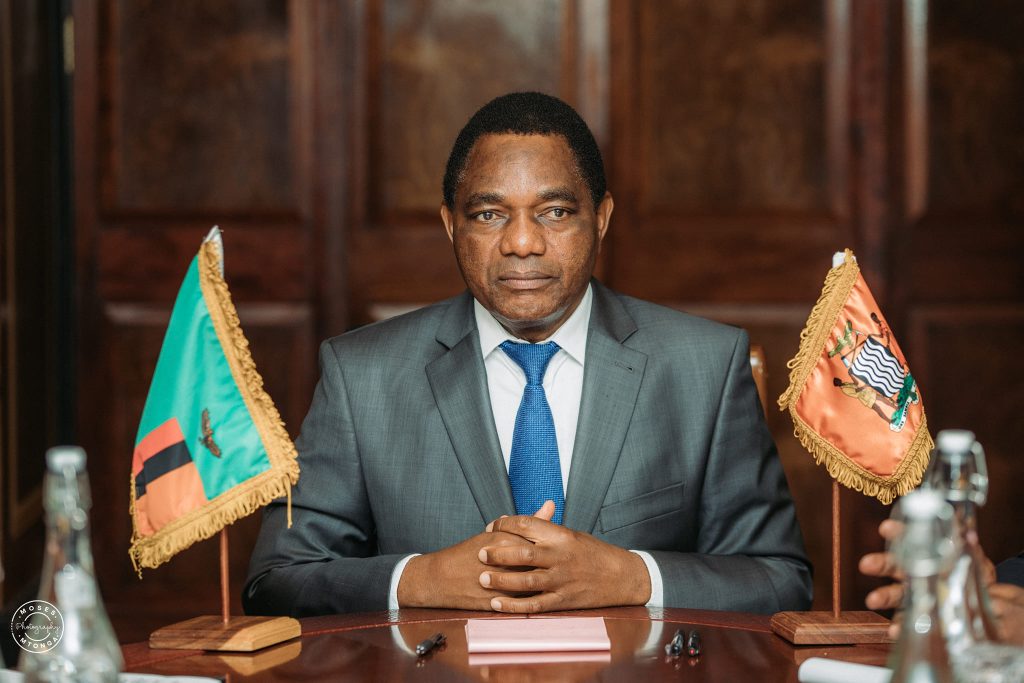 President Hakainde Hichilema seated at a table between the Zambian and presidential flags.