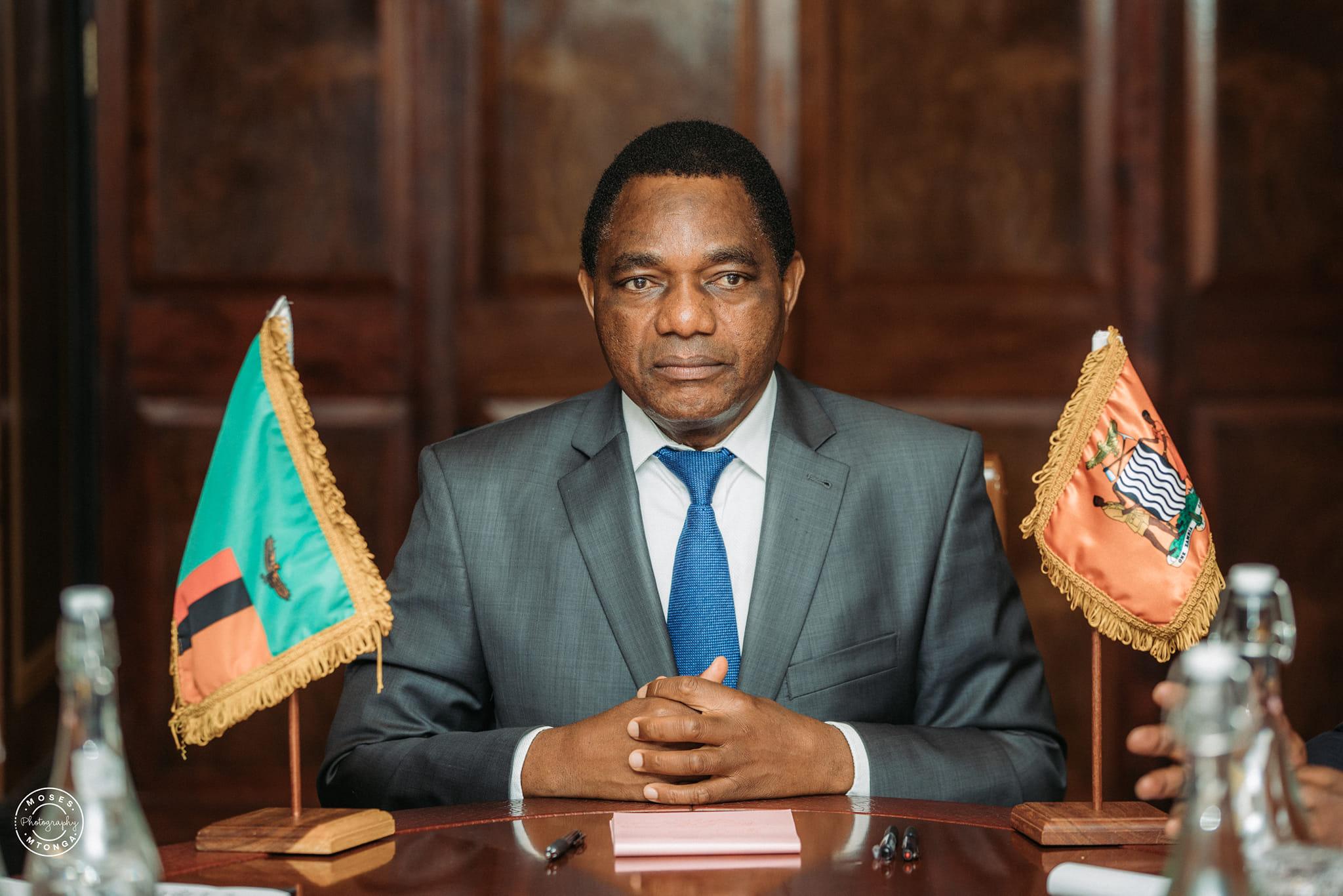 President Hakainde Hichilema seated at a table between the Zambian and presidential flags.