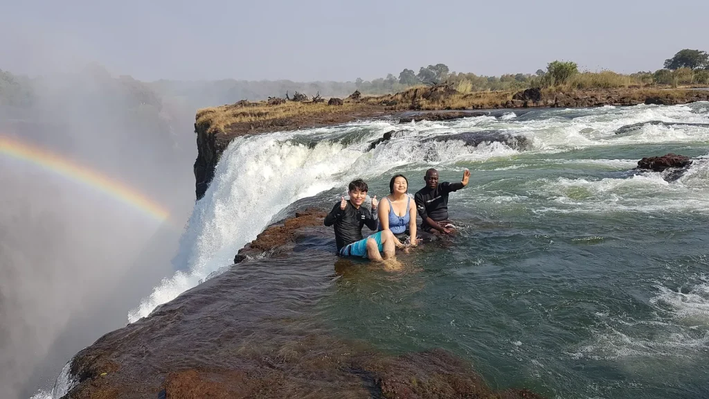 Visitors with a guide at Devil's Pool on the edge of the Victoria Falls on the Zambian side, with spray and a rainbow over the gorge