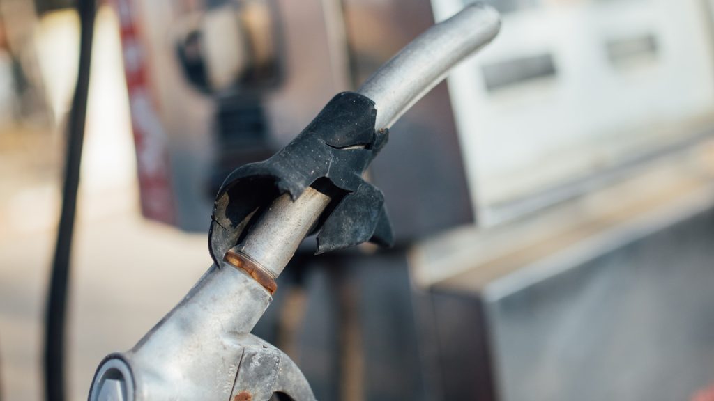 A hand holds a fuel nozzle beside a filling-station pump; illustrative photograph.