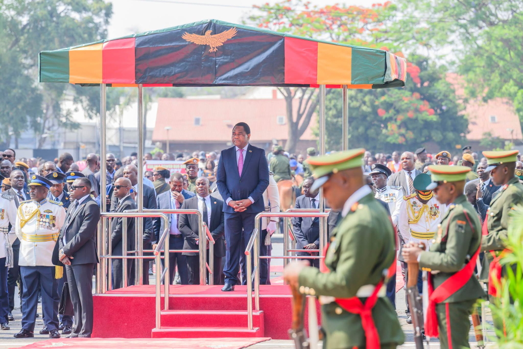 President Hakainde Hichilema stands beneath a canopy in Zambia’s national colours during a ceremony, with uniformed officers nearby.