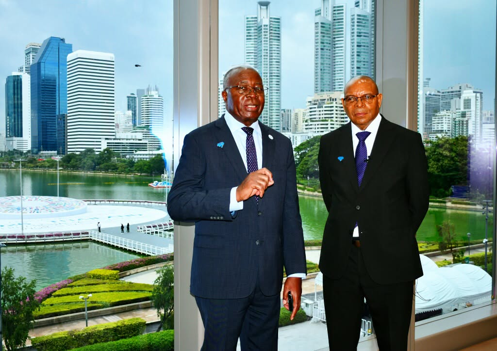 Two men in suits stand beside a window overlooking a lake and the Bangkok skyline.